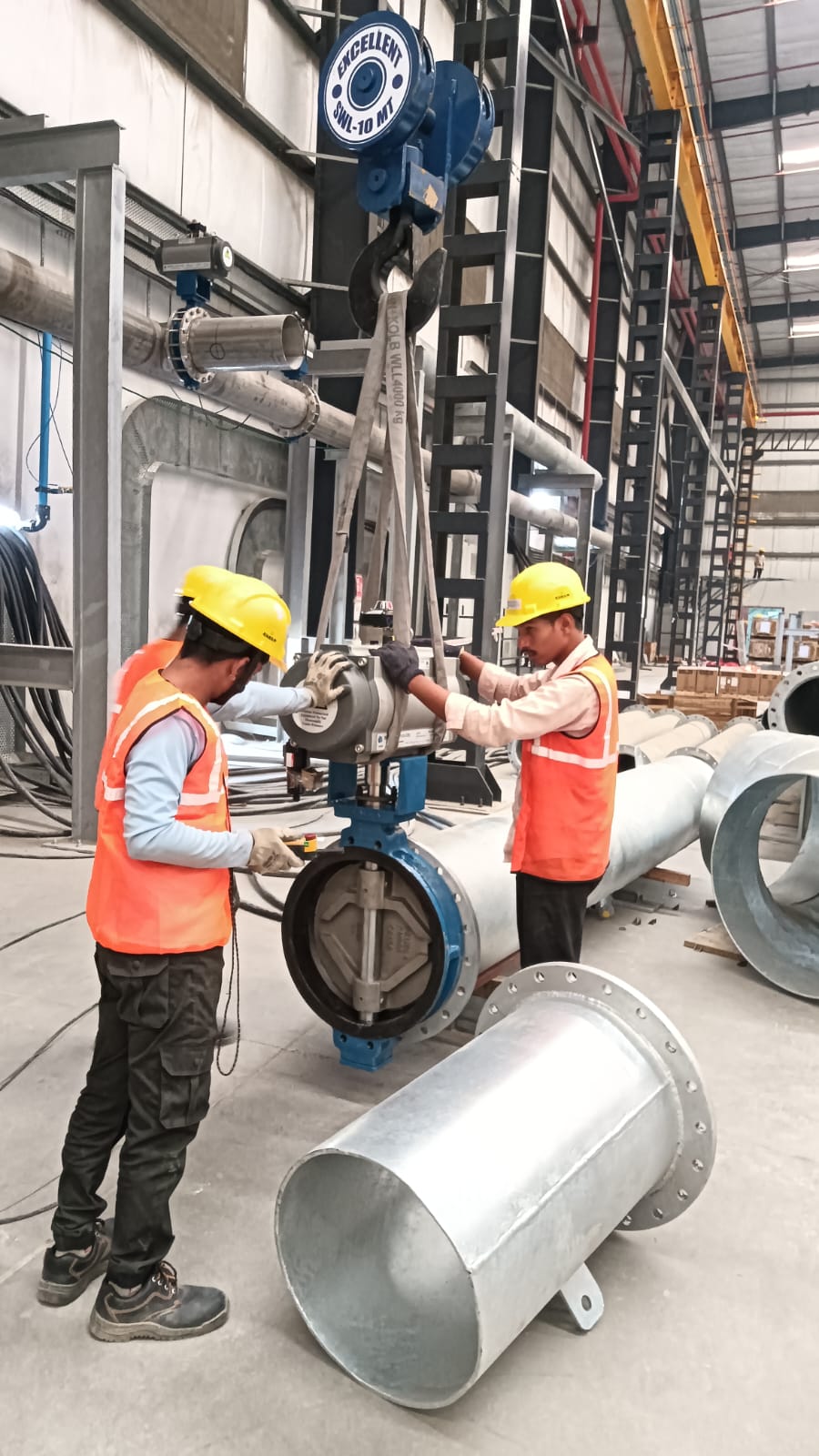 Workers fitting a large butterfly valve onto a flanged galvanized pipe spool in fabrication shop