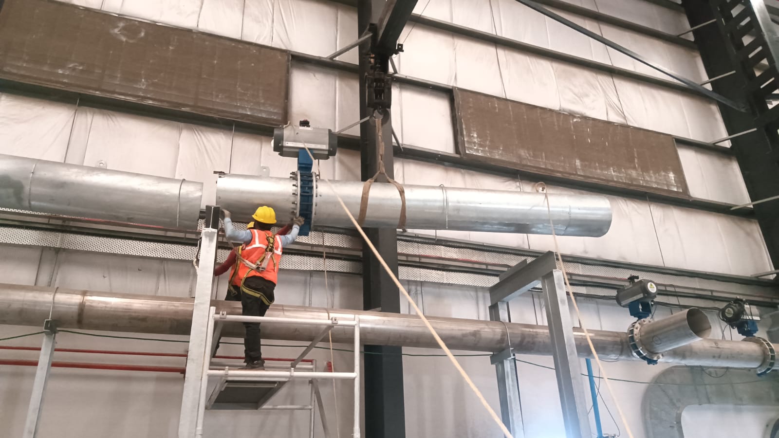 Worker on scaffold installing an actuated valve on an elevated overhead pipe run inside a PEB shed
